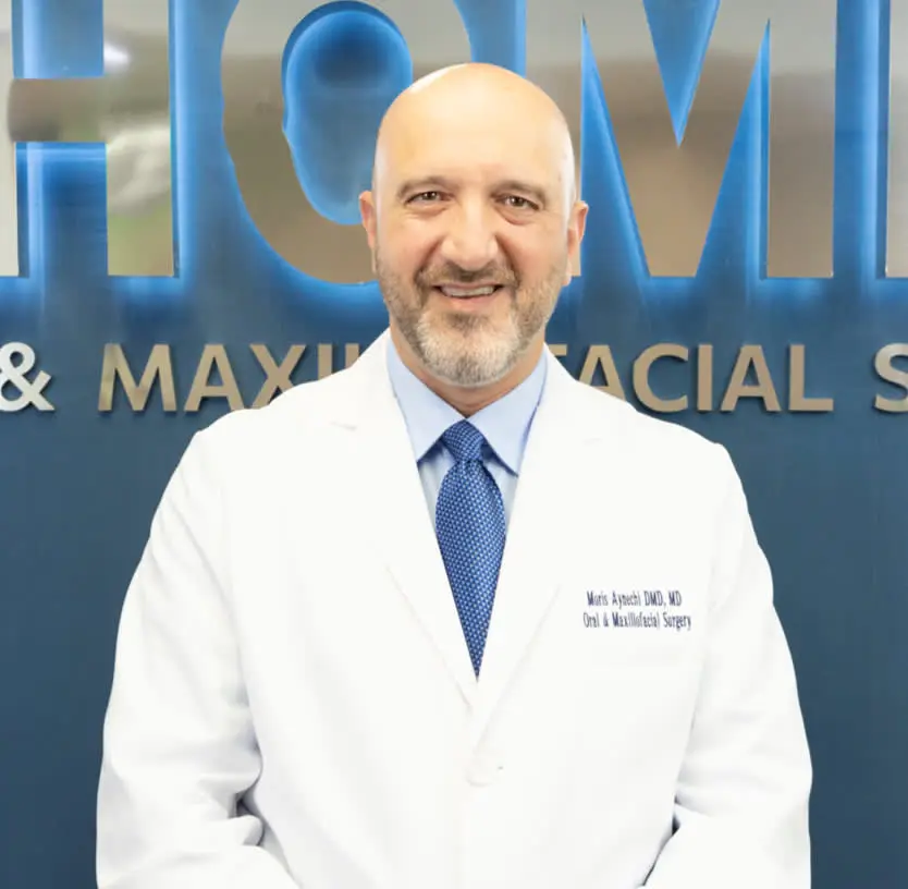 A person in a white coat and blue tie stands smiling in front of a sign.