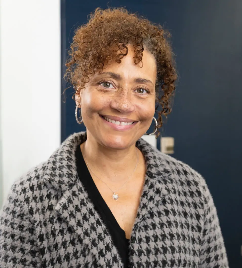 A person with curly hair smiling, wearing a houndstooth patterned coat, hoop earrings, and a necklace, standing indoors with a dark blue wall in the background.