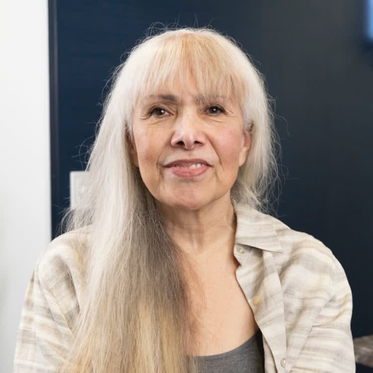 A woman with long gray hair smiles softly, wearing a plaid shirt over a gray top, against a dark background.