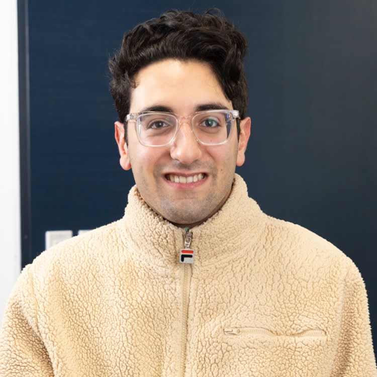 A person with glasses smiles, wearing a beige fleece jacket, standing in front of a dark blue background.