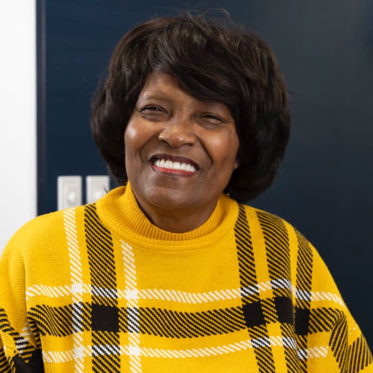 Woman smiling, wearing a yellow plaid sweater, against a dark background.