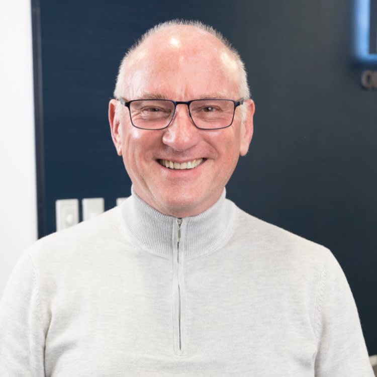 A man with glasses and a white sweater smiles in front of a dark background.