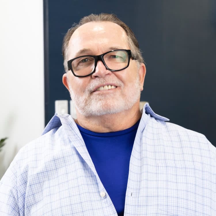 A man with glasses, wearing a light checked shirt over a blue top, smiling at the camera.