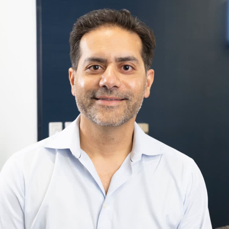 A man with short dark hair and a beard is smiling and wearing a light blue button-up shirt, standing indoors with a dark wall in the background.