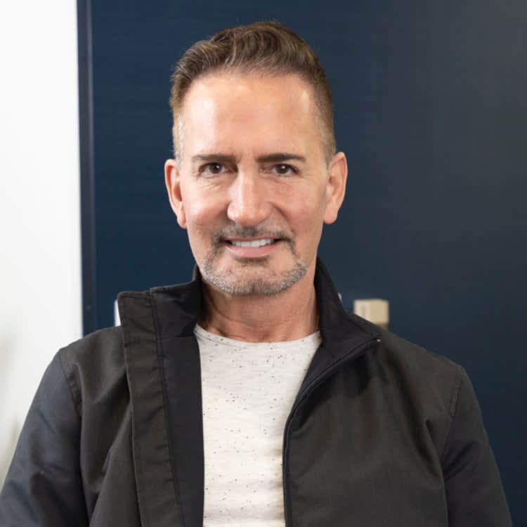 A person with short hair and a beard wearing a black jacket and white shirt, seated in front of a dark blue wall.