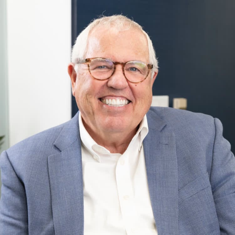 Smiling man with glasses and white hair in a blue suit jacket and white shirt, sitting indoors.